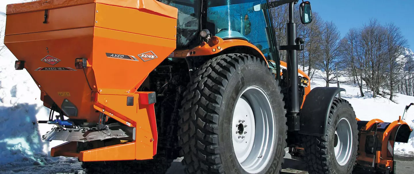 La spandiconcime, spargisale e spargisabbia AXEO al lavoro su una strada di montagna innevata con uno sciatore