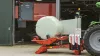 RW 1110 mentre fascia una balla tonda di fronte a una stalla con robot di alimentazione sullo sfondo.